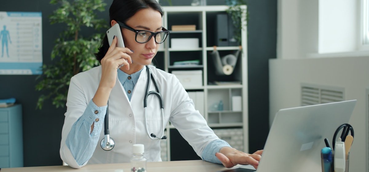 Physician in a clinic office reviewing billing on a laptop while taking a call
